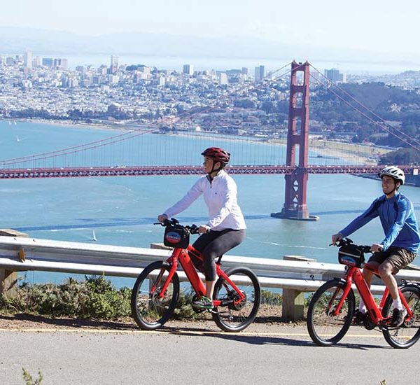 Two people riding tandem bike with San Francisco skyline in background